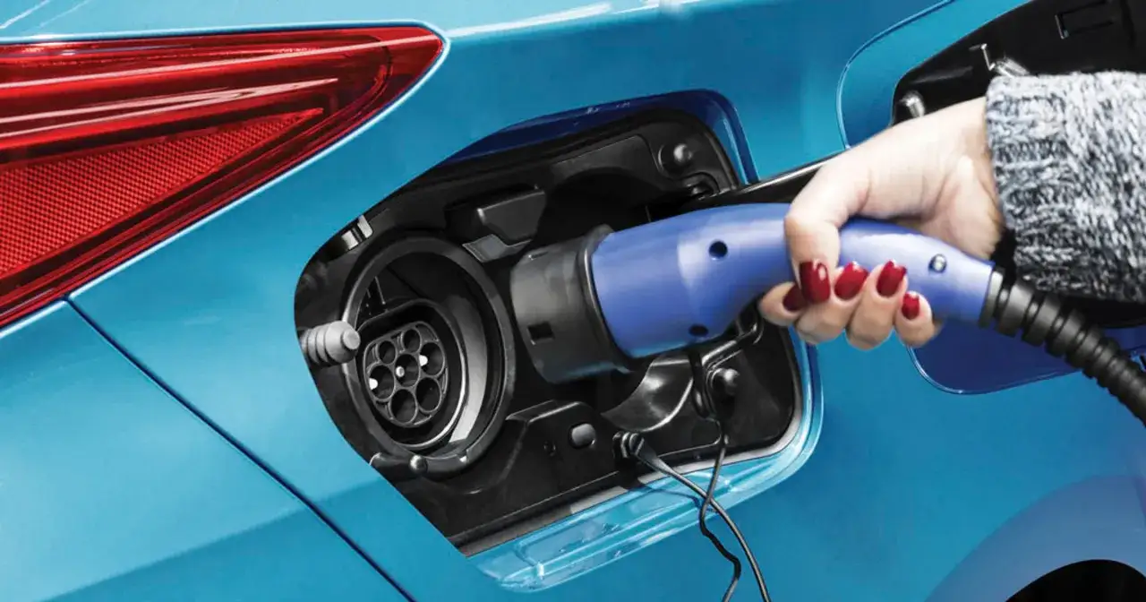 Dłoń z czerwonym lakierem na paznokciach wkłada niebieską wtyczkę do gniazda ładowania samochodu. To przyszłość motoryzacji – hybryda plug-in gotowa do jazdy.