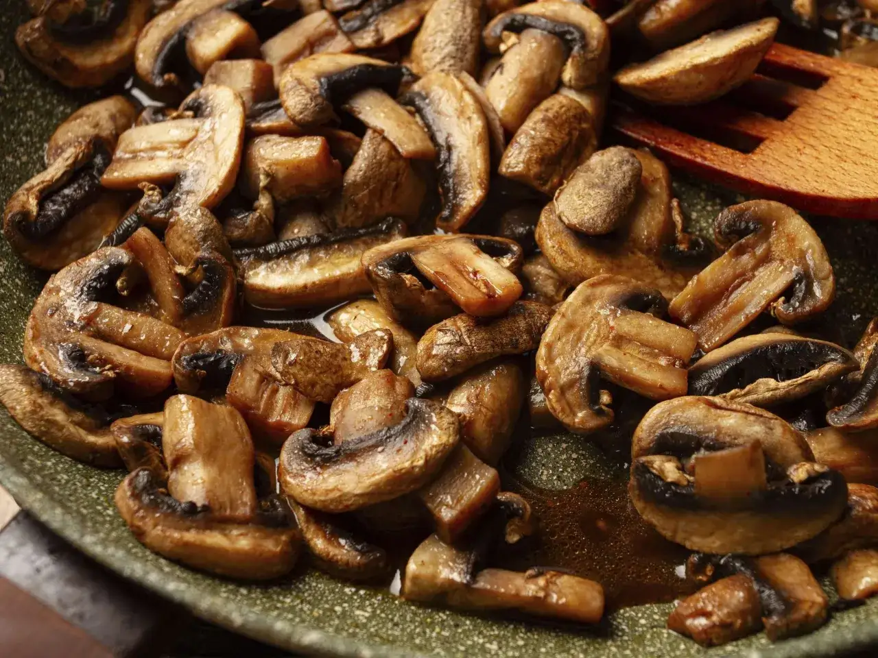 Gebratene Champignons in einer Pfanne, dampfend und glänzend, mit einem hölzernen Kochlöffel.