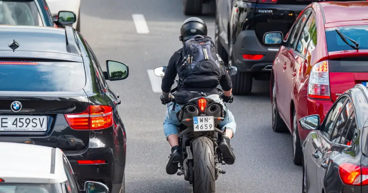 Motocyklista w kasku i plecaku przeciska się między samochodami w korku. Widać znaki motocyklistów na tablicy rejestracyjnej.