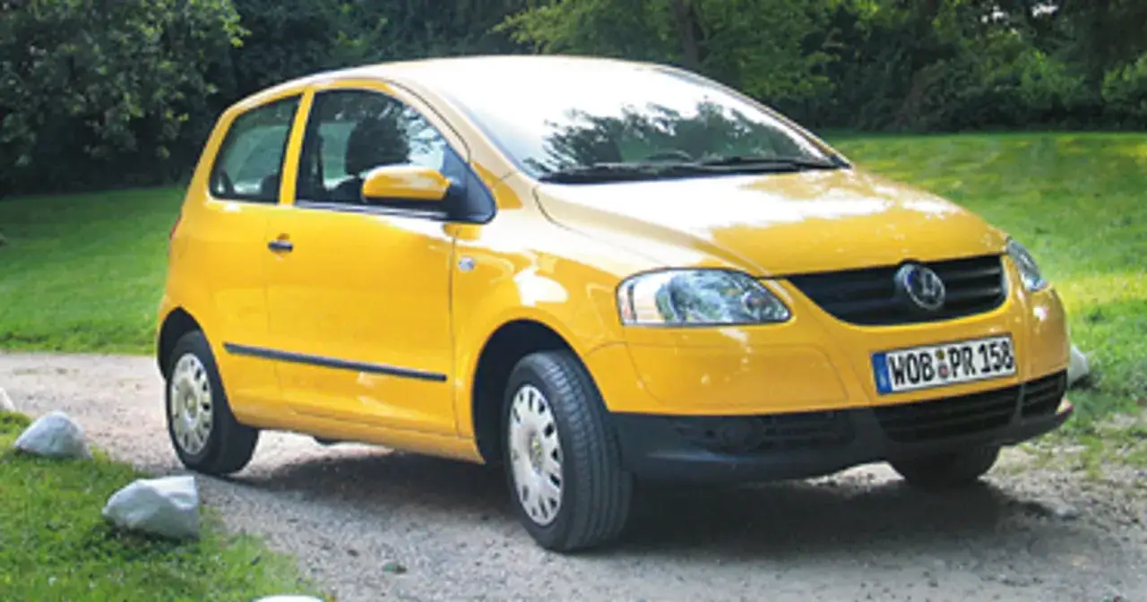 Żółty Volkswagen Fox zaparkowany na żwirowej drodze, otoczony zielenią.