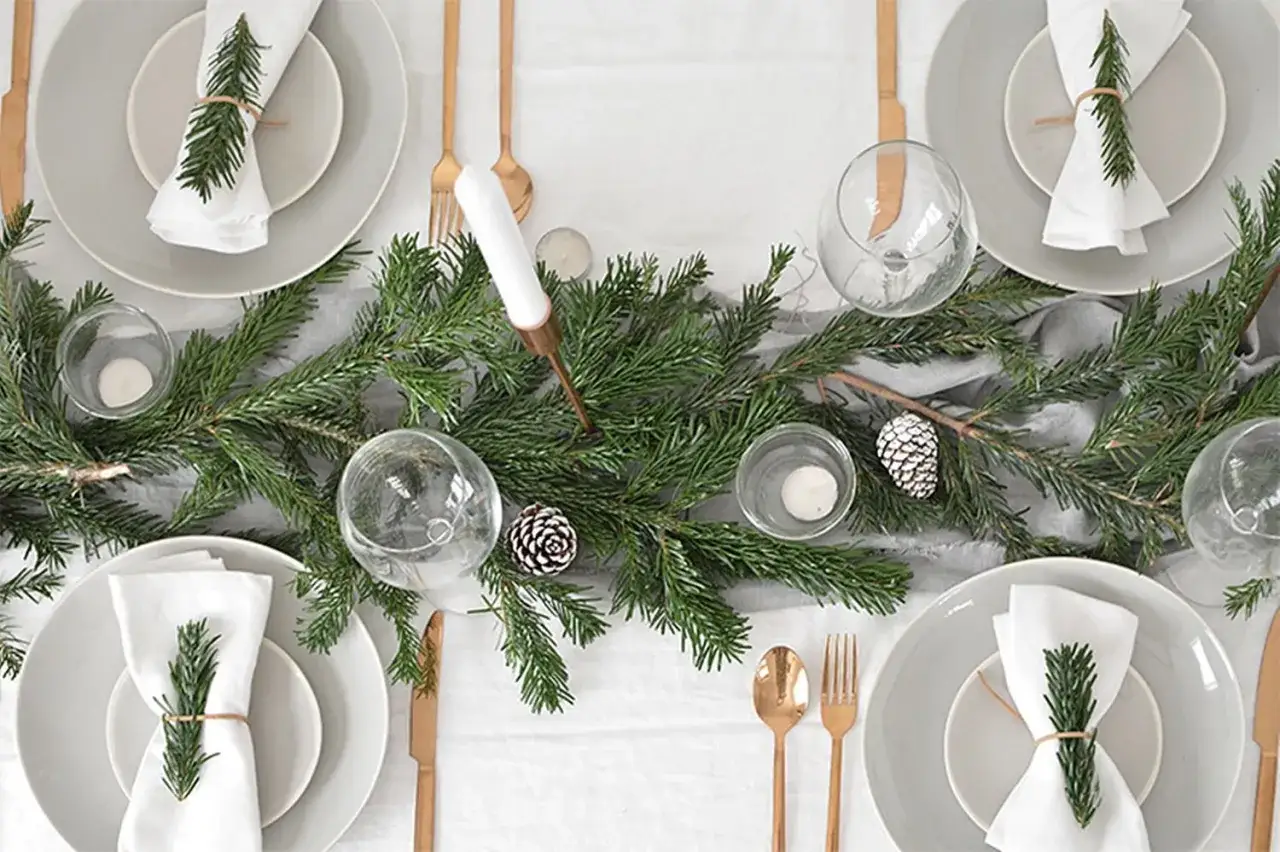 Sencillo decoracion mesa navidad con ramas de pino, piñas y velas. Vajilla gris, servilletas blancas con detalle de romero y cubertería dorada.