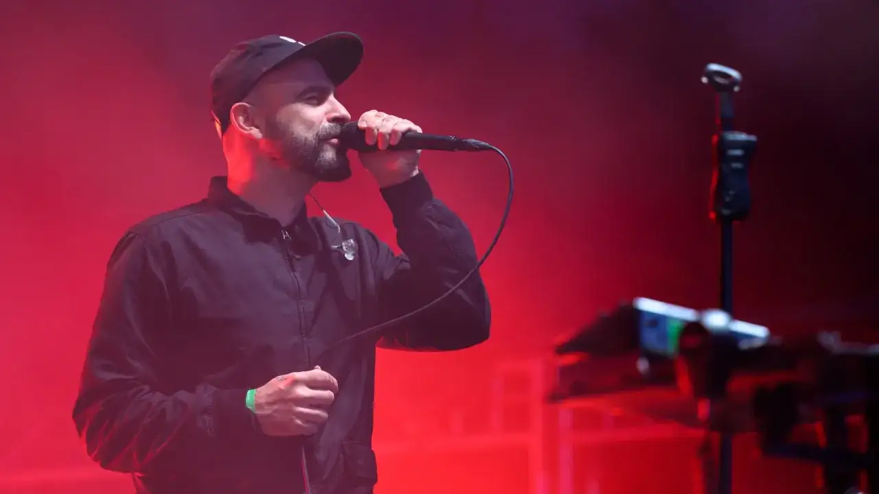 Wokalista z brodą i czapką na głowie śpiewa do mikrofonu na scenie. Czerwone światła tworzą atmosferę koncertu. Tauron Nowa Muzyka line-up.