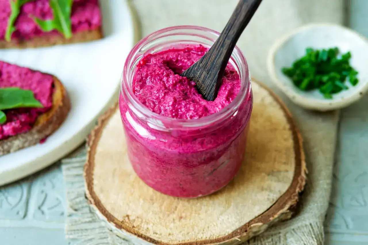 Glas mit cremigem, veganem Rote Bete Aufstrich, daneben Brot mit Aufstrich und Schnittlauch.