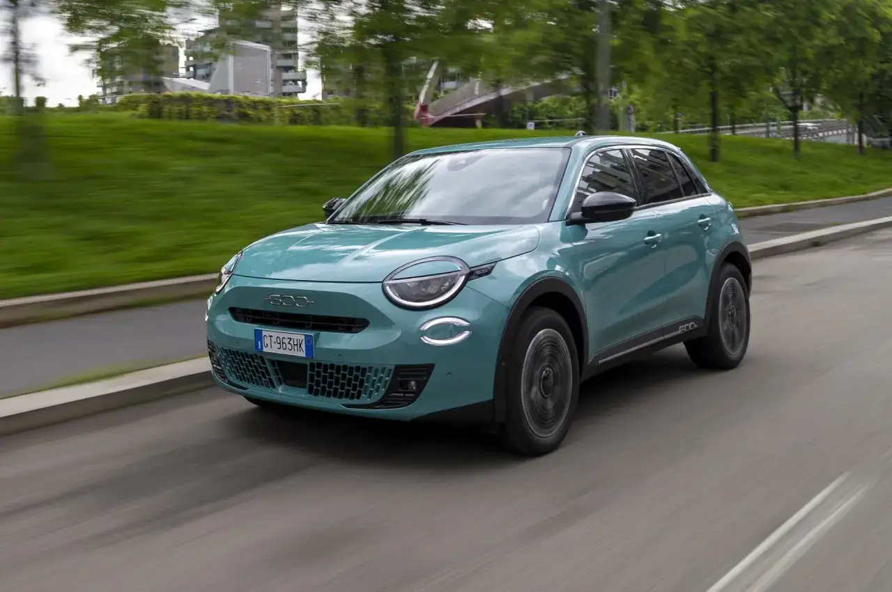 Nowoczesny, turkusowy Fiat 600 Hybrid sunie po miejskiej drodze, otoczony zielenią i budynkami.
