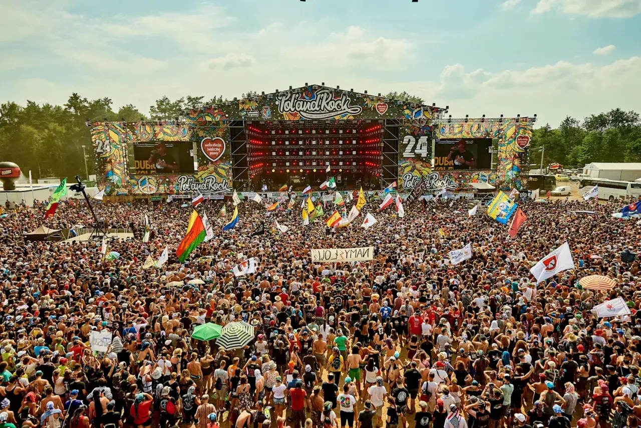Tłum ludzi na festiwalu Poland Rock, gdzie jest scena i ekrany. Wszędzie flagi i słońce.