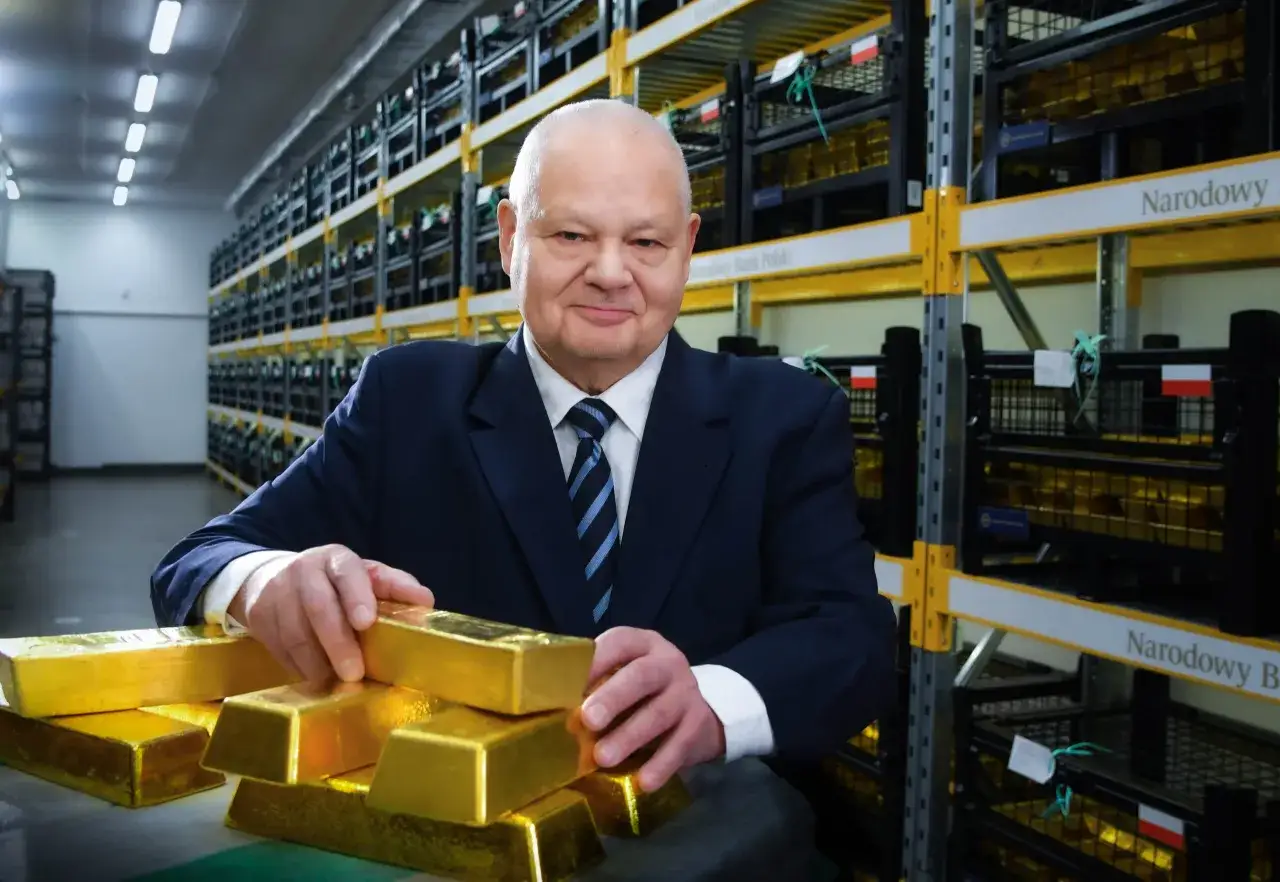 Mężczyzna w garniturze w magazynie Narodowego Banku Polskiego pokazuje sztabki złota, sugerując, ile złota ma Polska.