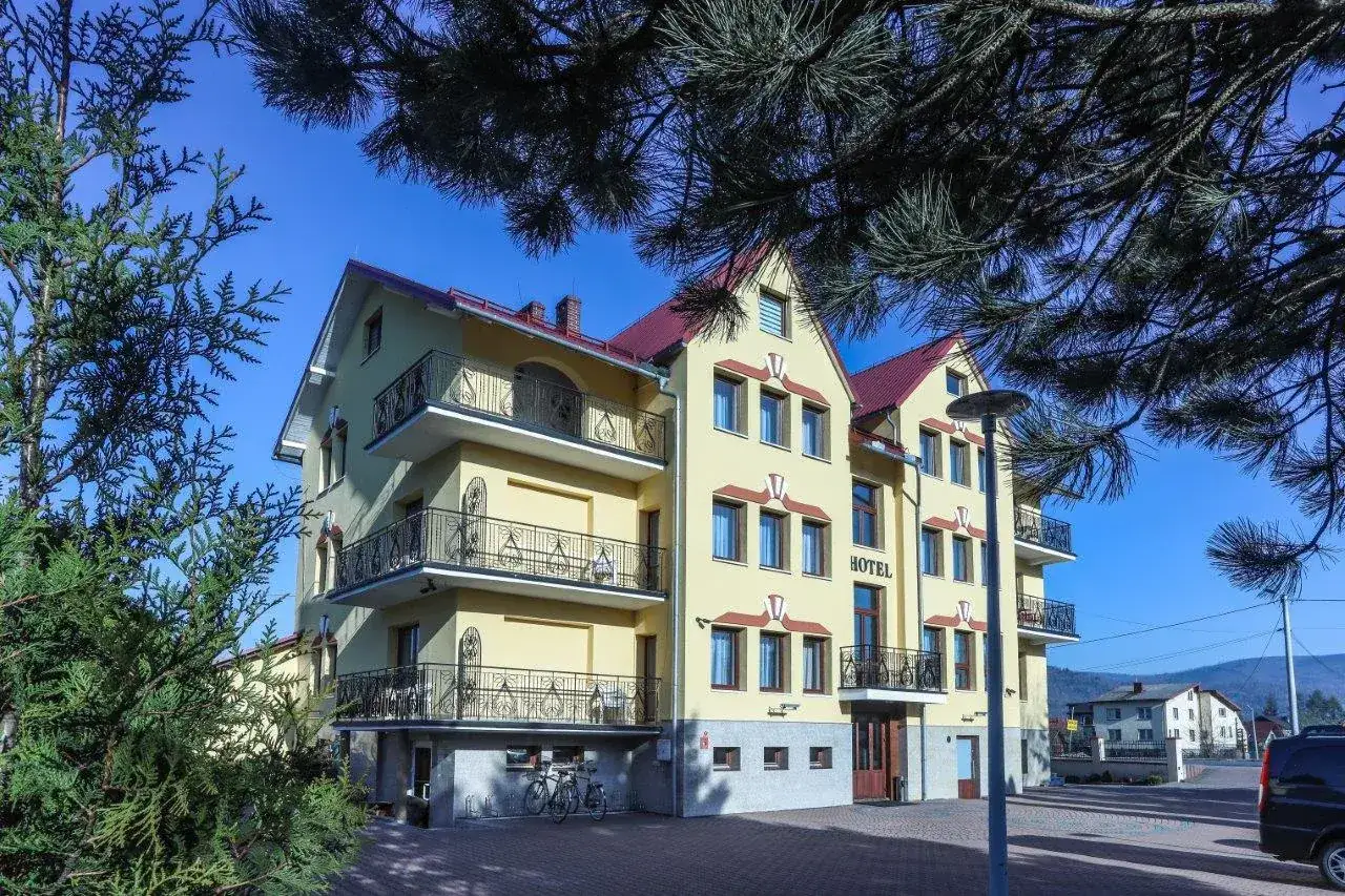 Hotel Milena w Milówce, żółty budynek z balkonami i czerwonym dachem, otoczony zielenią.