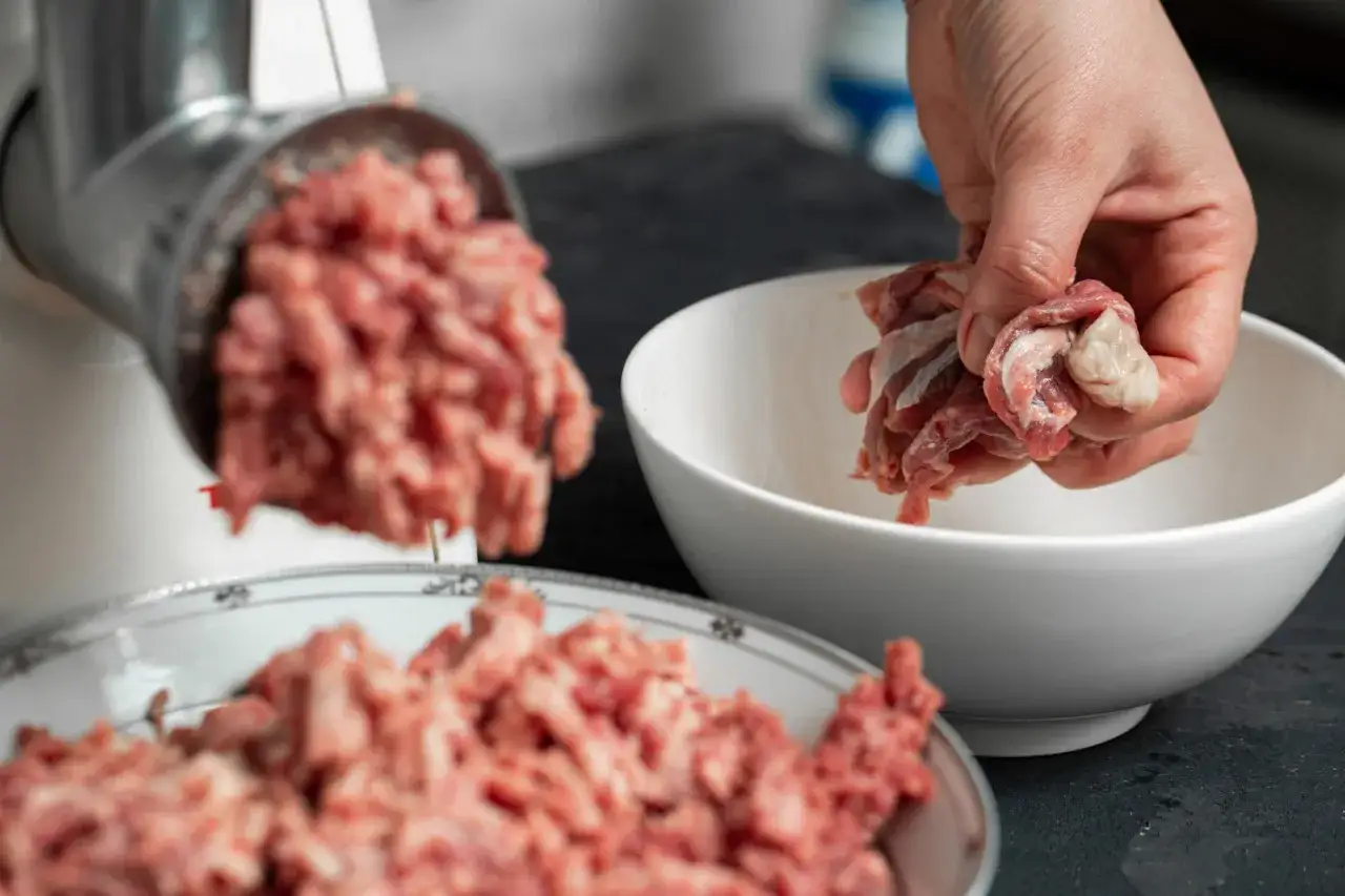 Ręka wkłada mięso do maszynki, przygotowując je do mielenia. Czy pies może jeść serca drobiowe? To mięso może być składnikiem jego diety.