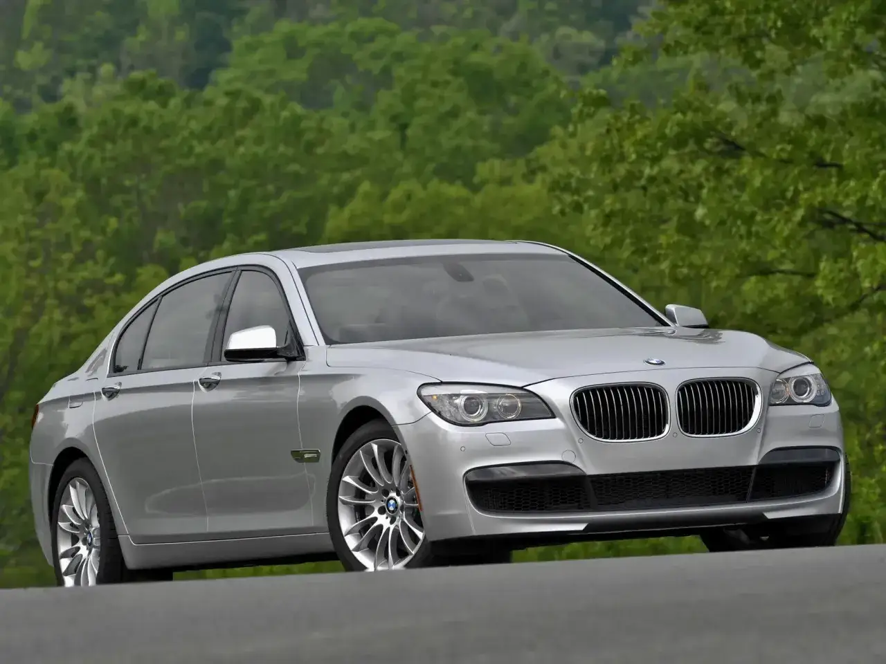 Srebrne BMW 7 F01 na tle zielonych drzew. Elegancka limuzyna prezentuje się okazale na drodze.