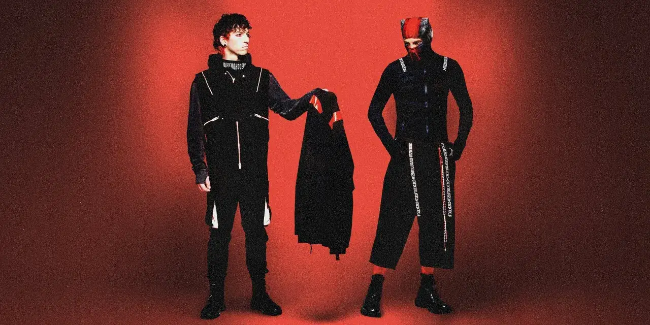 Dwóch artystów w czarnych strojach, przypominających styl z okładek płyt twenty one pilots, pozuje na tle czerwonym.