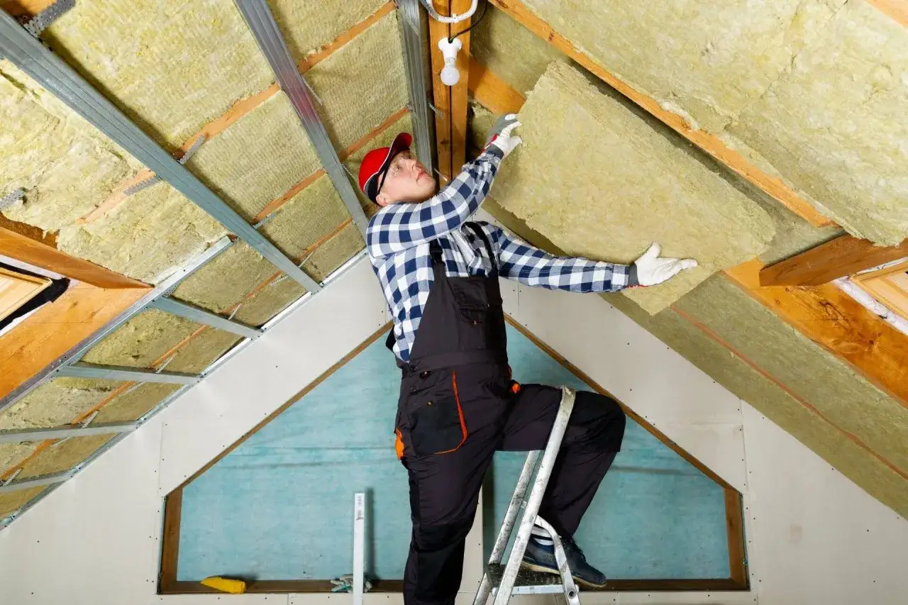 Pracownik na drabinie układa wełnę mineralną na poddaszu. To kluczowy etap, by wiedzieć, jak uszczelnić właz na strych i zapewnić ciepło.