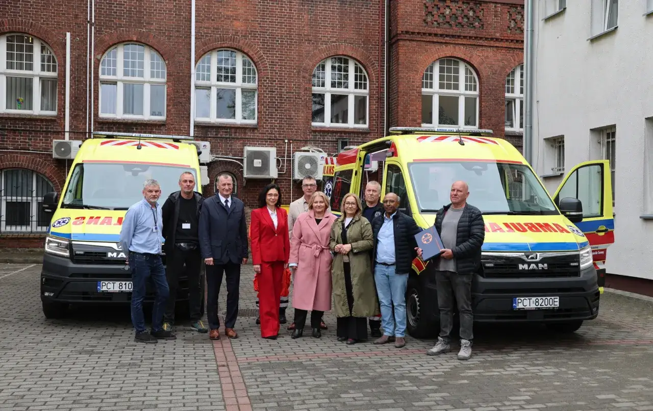 Grupa osób pozuje przed dwoma ambulansami. Jeden z mężczyzn trzyma dokument, być może związany z czn rejestracja.