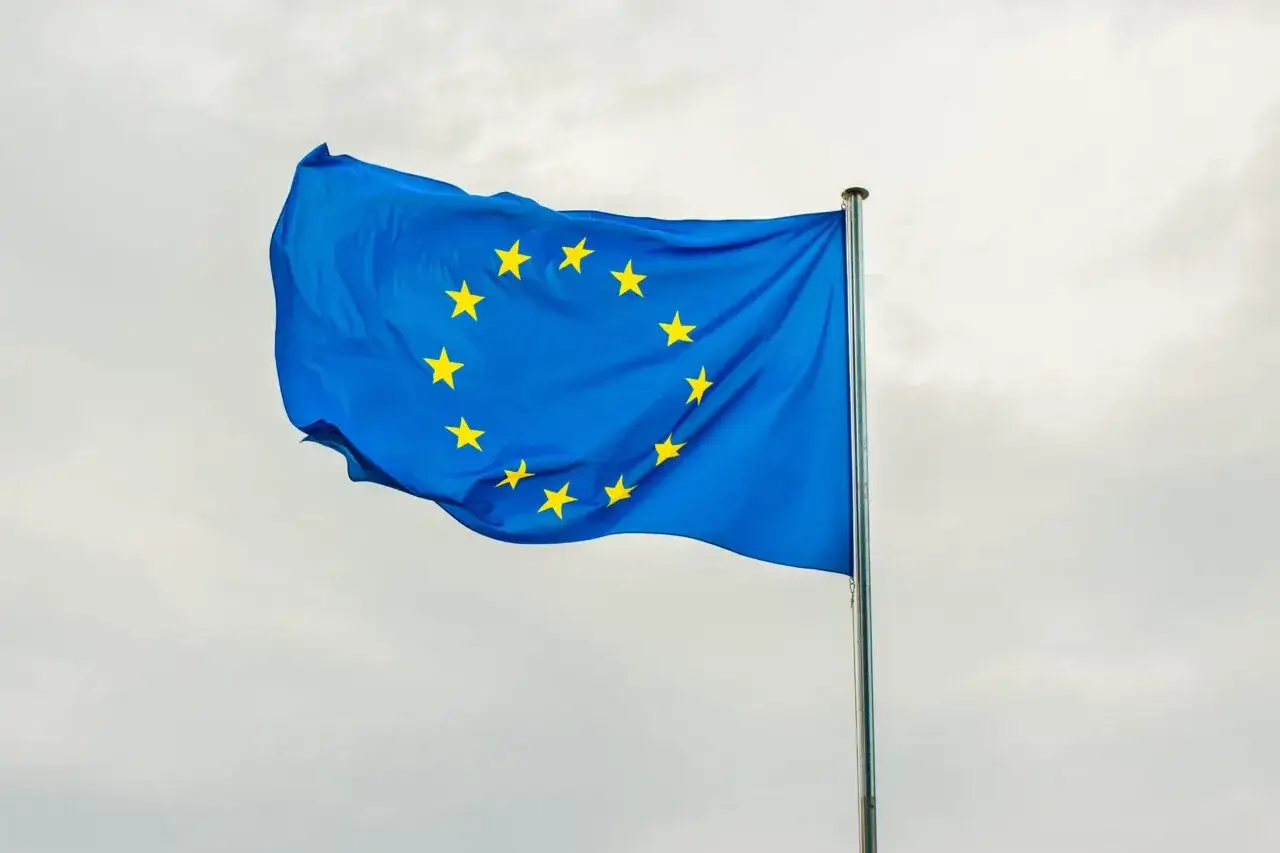 Niebieska flaga z żółtymi gwiazdami, symbolizująca europejski tytuł egzekucyjny, powiewa na maszcie na tle pochmurnego nieba.