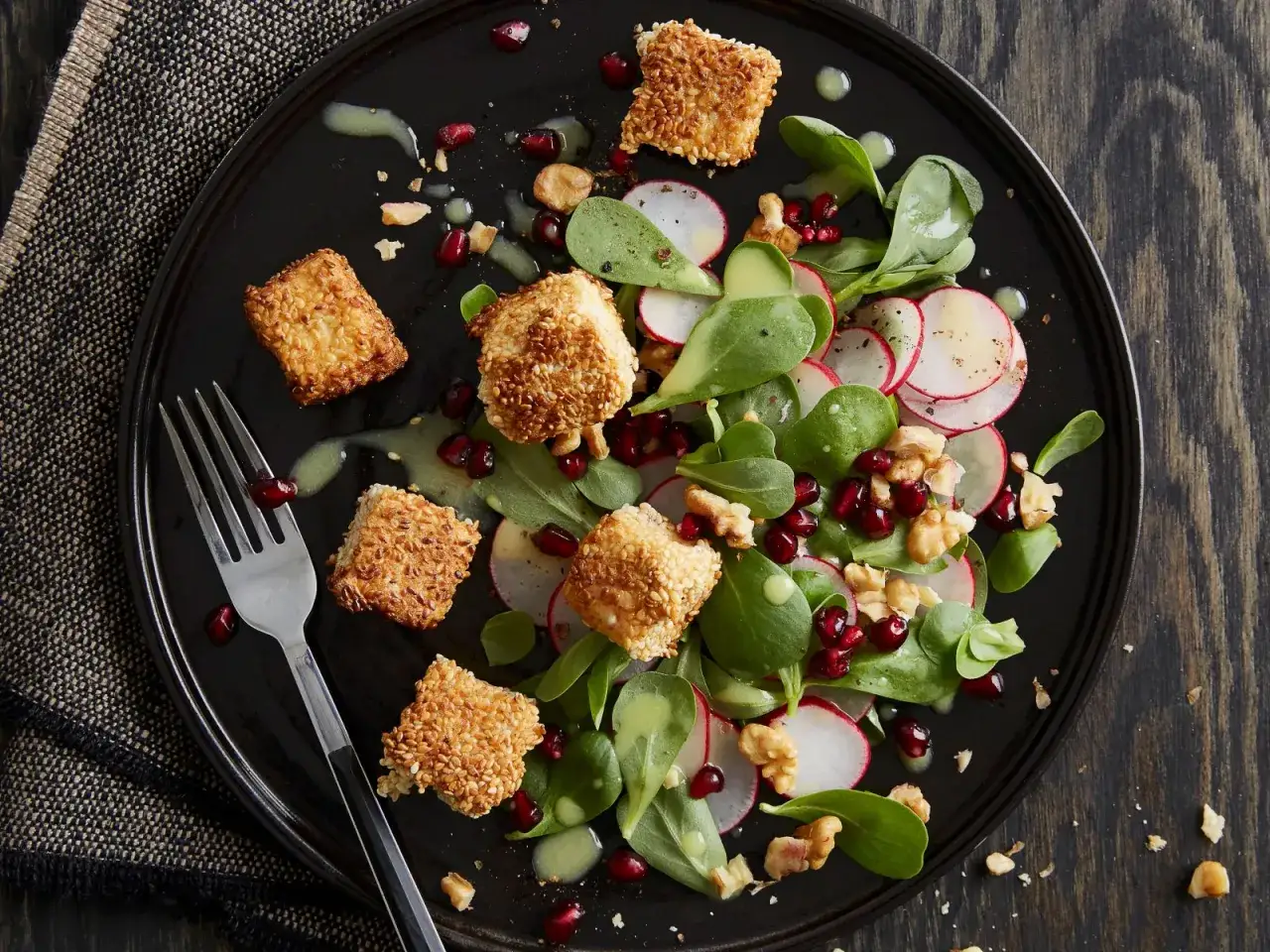 Feldsalat mit Granatapfelkernen, Radieschen und Sesam-Käsewürfeln auf schwarzer Platte.