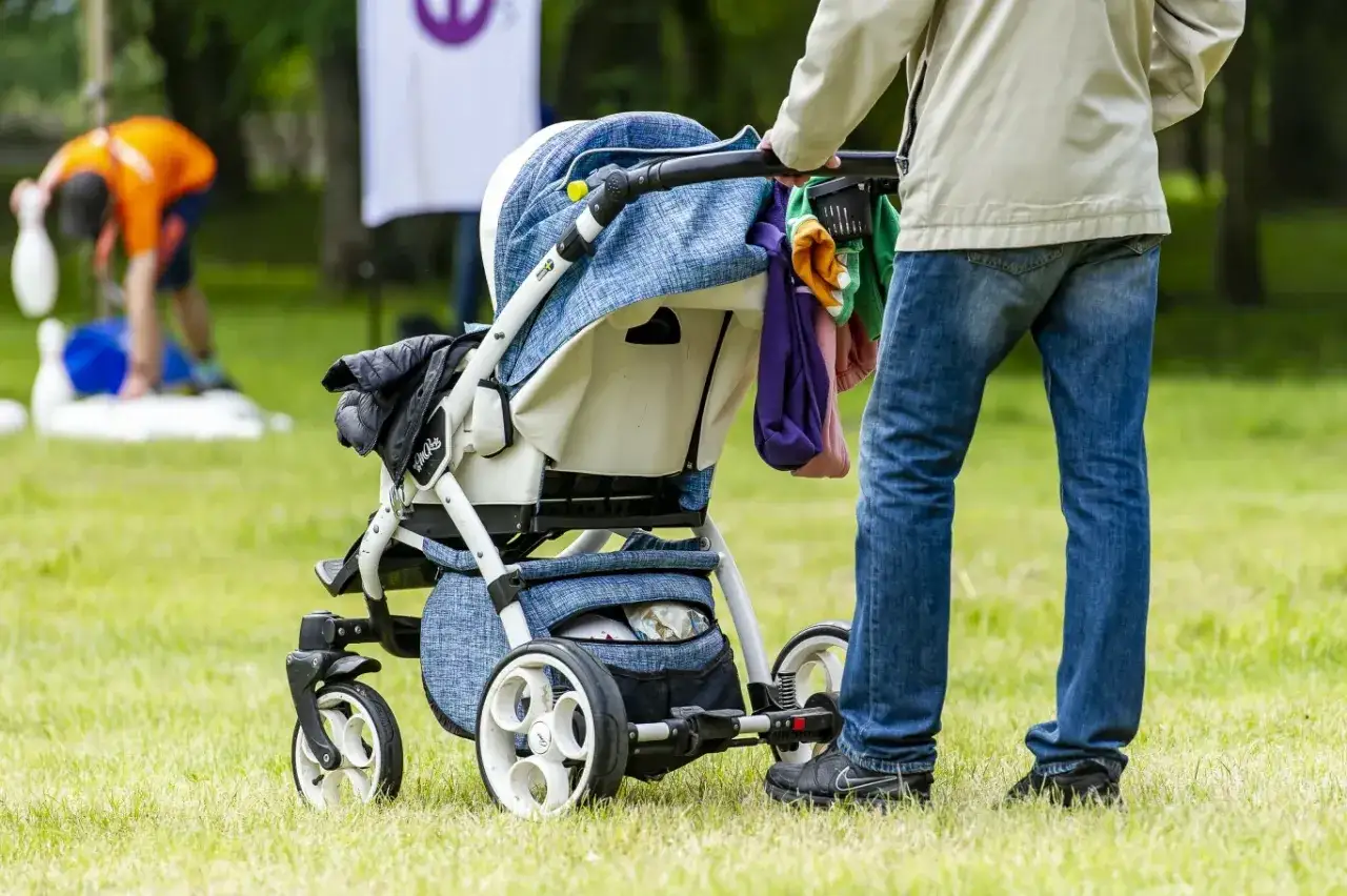 Osoba na urlopie rodzicielskim pcha wózek dziecięcy po trawie. W tle widać bawiących się ludzi.