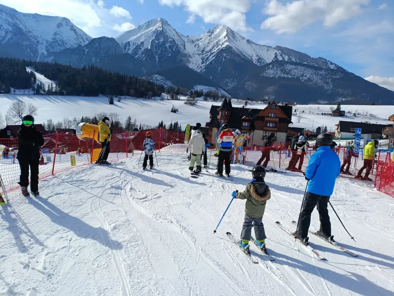 Ludzie na nartach i snowboardach w słoneczny dzień w ski centrum Strachan, z majestatycznymi ośnieżonymi górami w tle.
