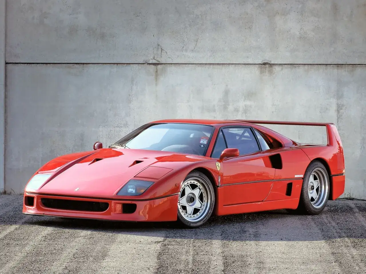 Ikoniczny czerwony Ferrari F40 z charakterystycznym skrzydłem, zaparkowany na tle betonowej ściany.