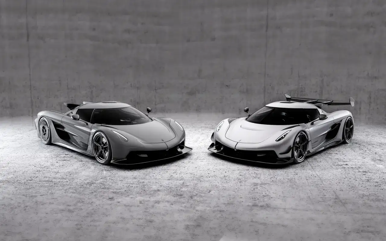 Dwa lśniące Koenigsegg Jesko, jeden szary, drugi srebrny, stoją obok siebie na betonowym tle.