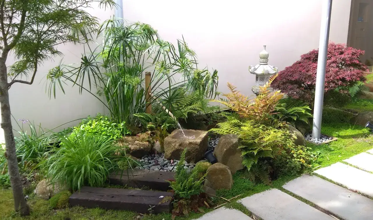 Un sereno mini jardín japonés en casa, con cascada de agua, rocas, plantas exuberantes y una linterna de piedra.