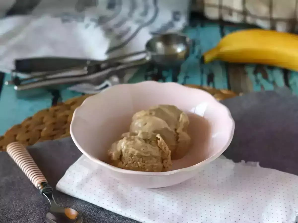 Zwei Kugeln cremiges, veganes Bananeneis in einer rosa Schale, daneben ein Löffel und ein Stück Gebäck. Eine Banane liegt im Hintergrund.