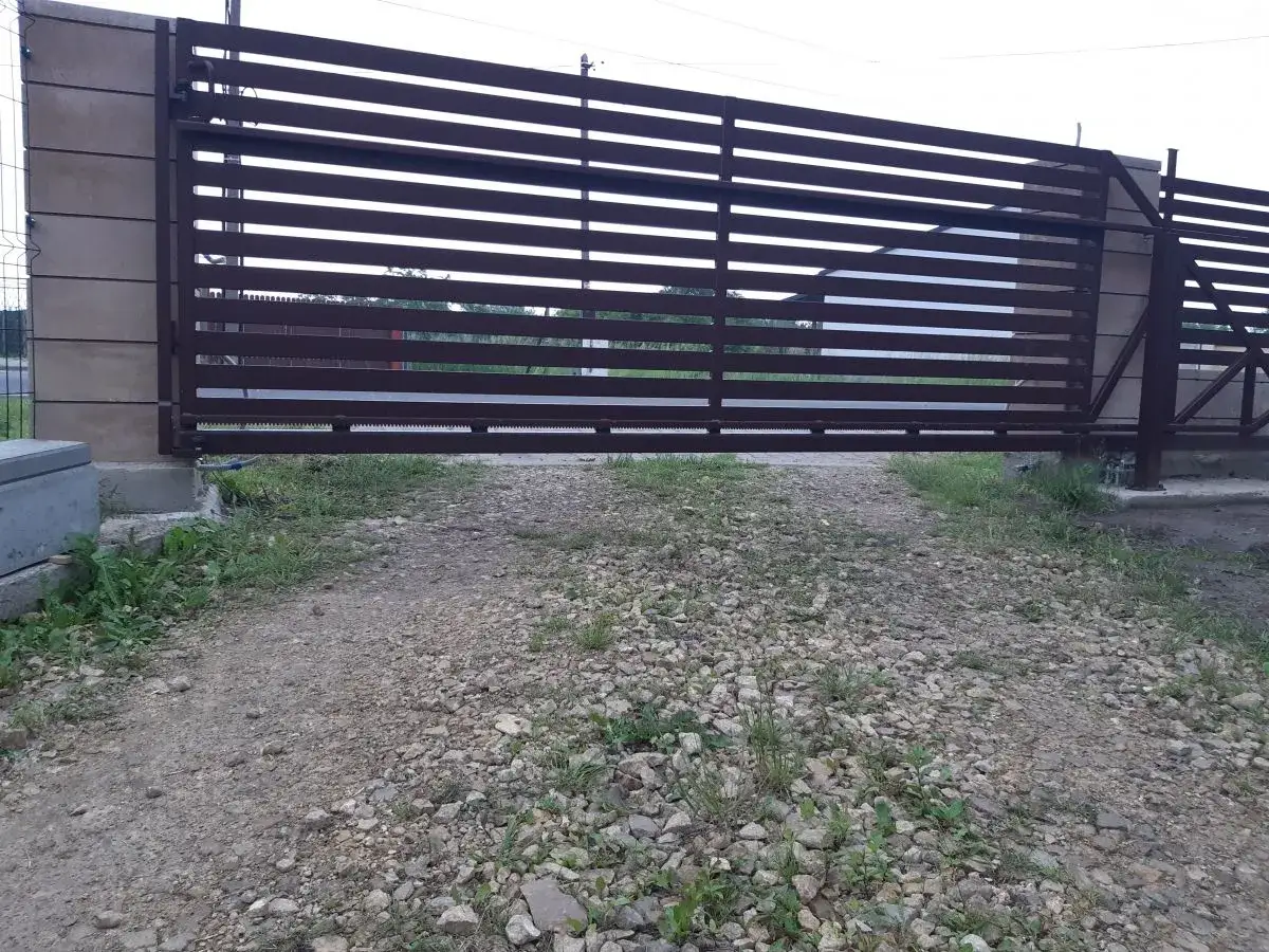 Brązowa, metalowa brama przesuwna z poziomymi szczeblinami. Widoczna jest szczelina pod bramą, przez którą widać trawę i żwir.