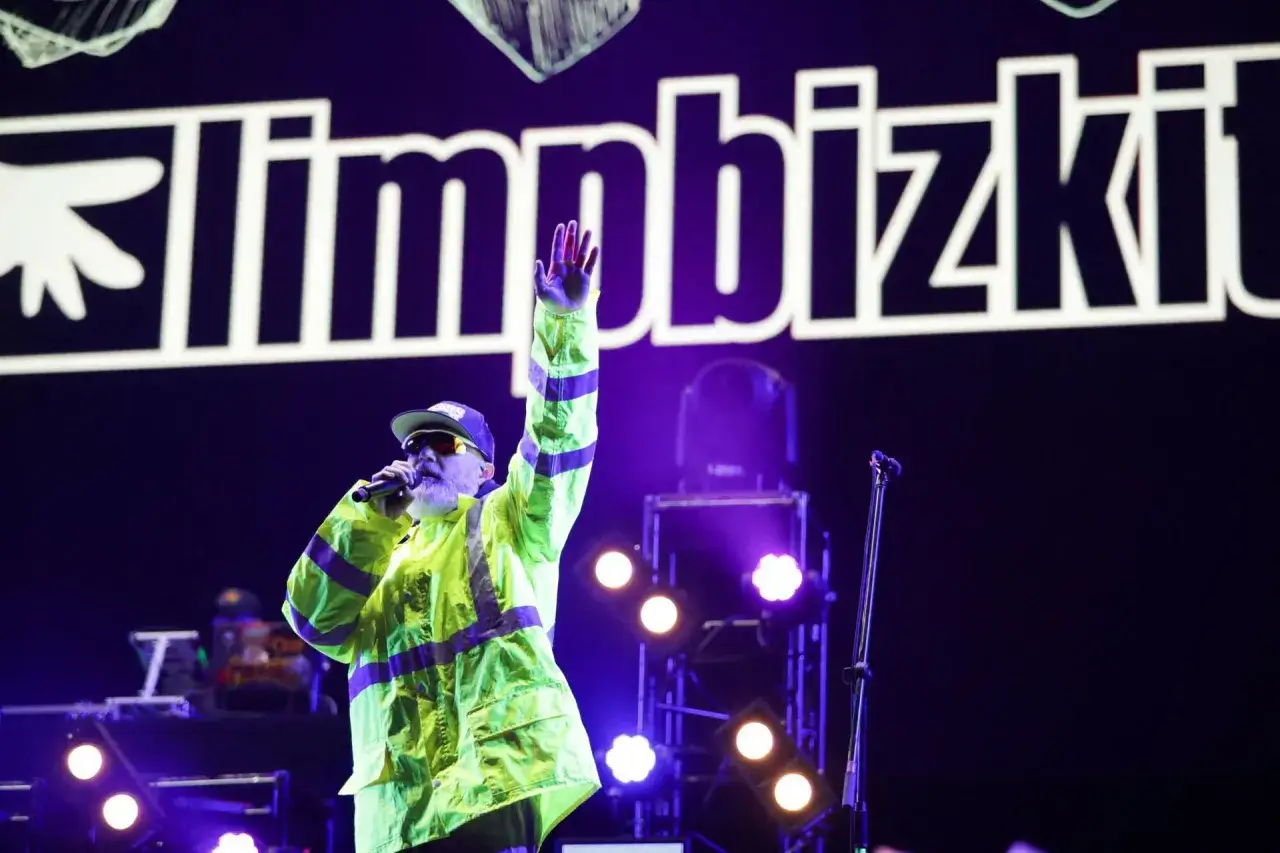 Fred Durst z Limp Bizkit na koncercie, w neonowej kurtce, z ręką w górze. Tło to logo zespołu.
