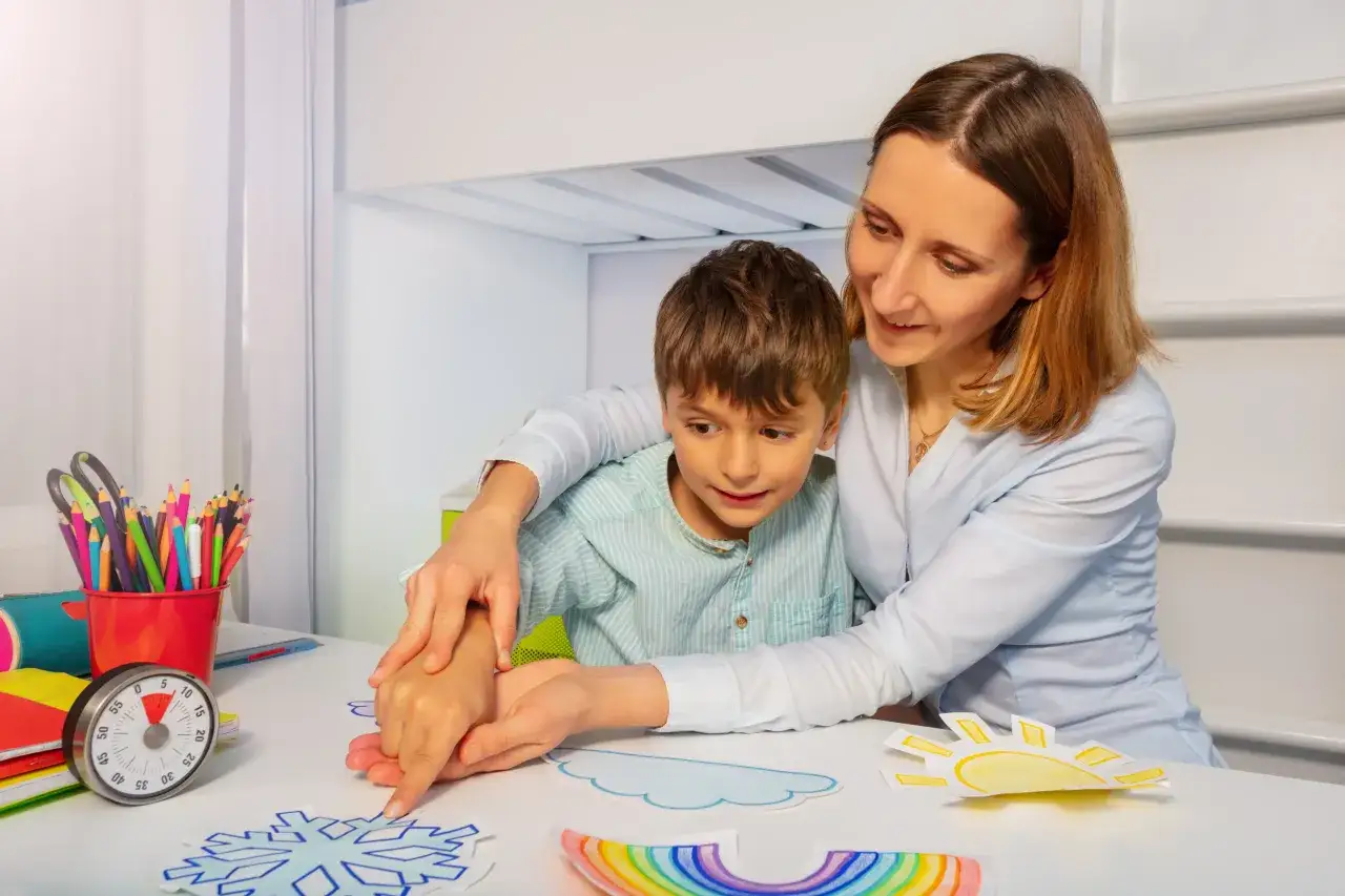 Kobieta pomaga chłopcu w zajęciach plastycznych, tworząc prace na temat pogody. To przykład wsparcia, które może być częścią świadczenia pielęgnacyjnego 2026.