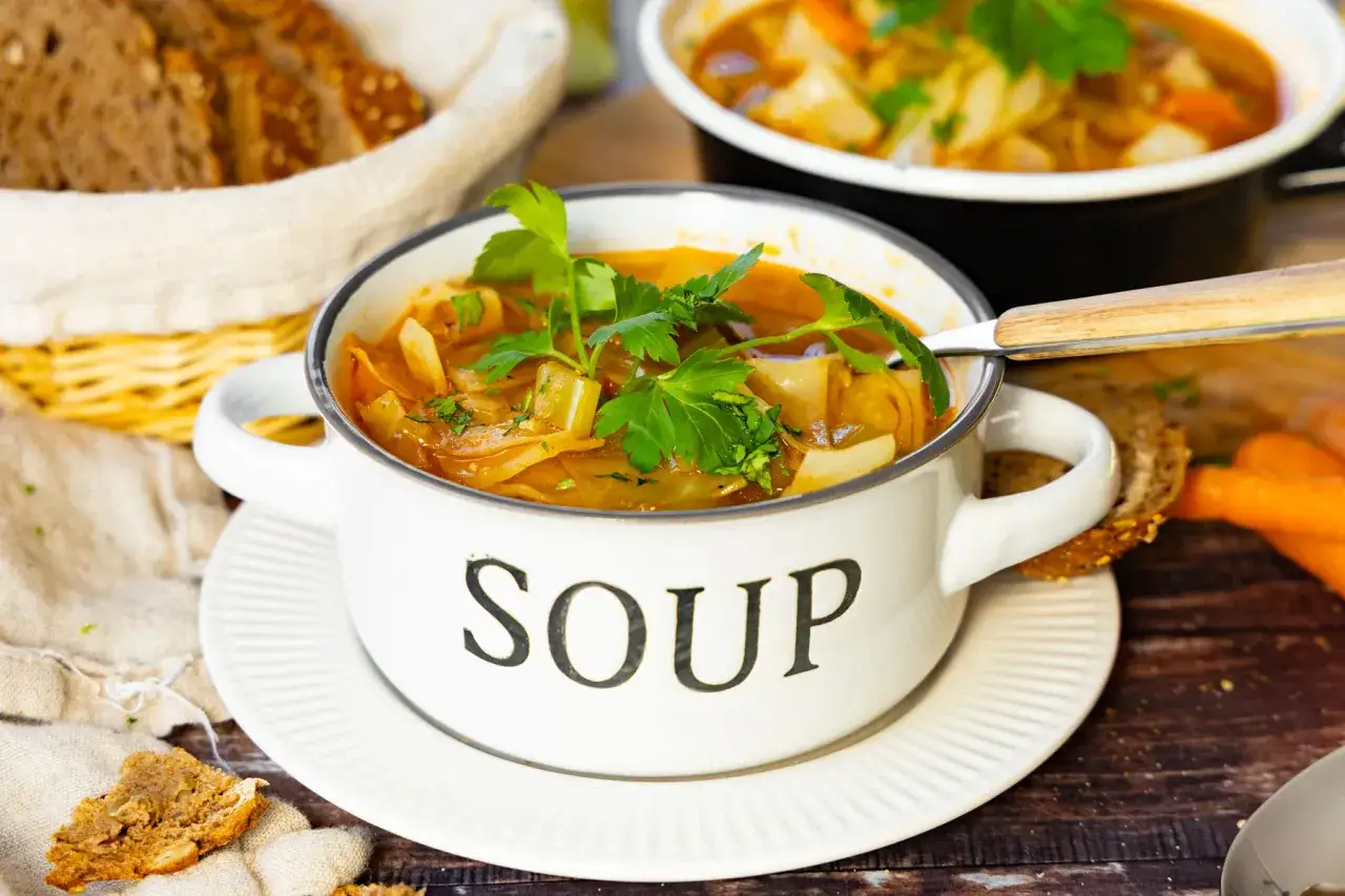 Herzhafte Kohlsuppe, low carb, in einem weißen Topf mit der Aufschrift "SOUP", garniert mit Petersilie. Dazu gibt es Vollkornbrot und Karotten.