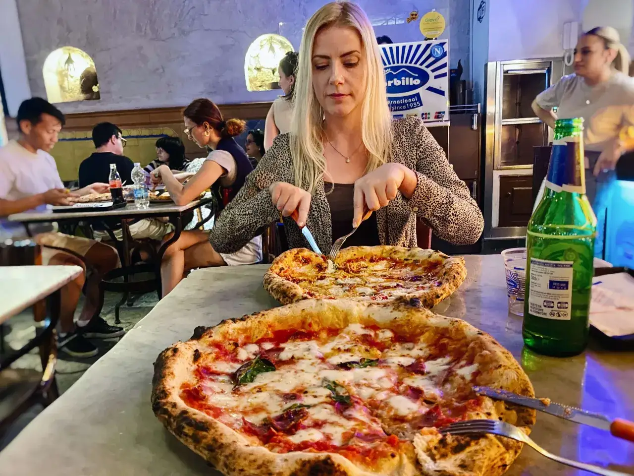 Kobieta kroi pizzę w restauracji, w tle inni ludzie i butelka napoju.