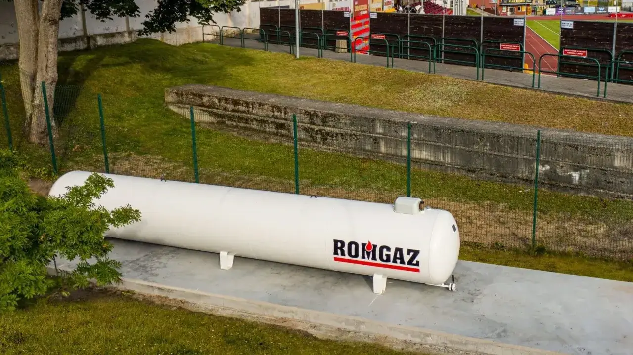 Biały zbiornik gazu ROMGAZ stoi na betonowej podstawie. Widać ogrodzenie i trawę, a w tle trybuny stadionu. Odległość gazociągu od ogrodzenia jest widoczna.
