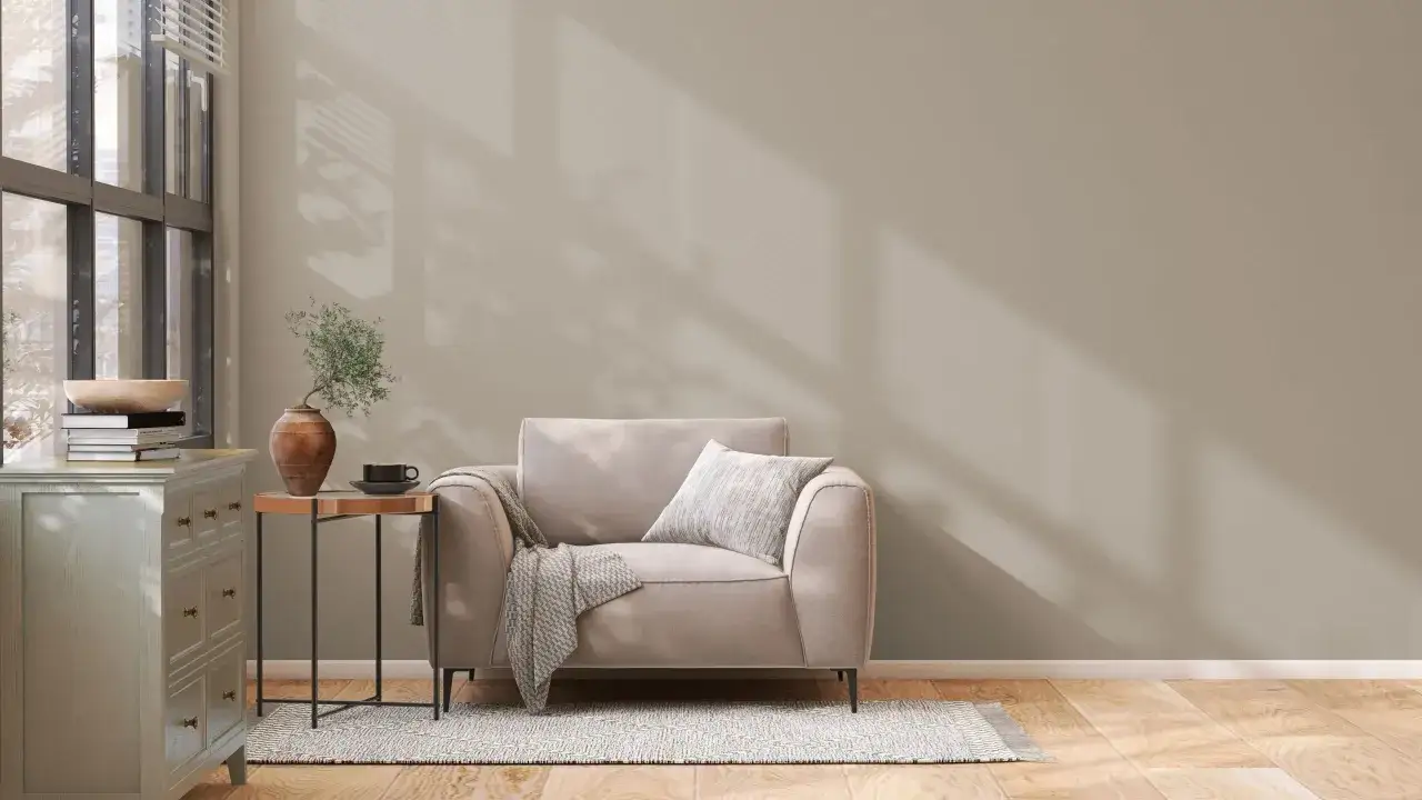 Un sillón beige con manta y cojín, junto a una mesita auxiliar con jarrón y taza. La pared tiene unos colores de pintura neutros, con sombras de luz.