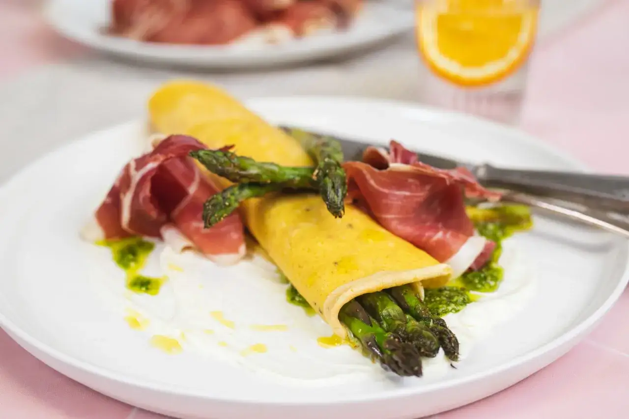 Ein Pfannkuchen mit Spargel, umhüllt von zartem Schinken, serviert auf einer cremigen Basis mit Kräutersauce.