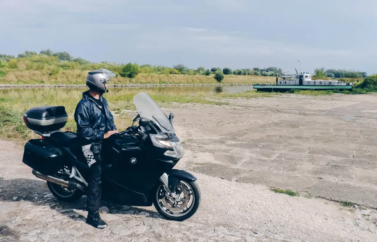 Motocyklista w kasku i kurtce stoi obok czarnego motocykla nad kanałem z łodzią w tle.