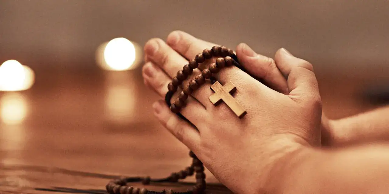 Dłonie trzymające drewniany różaniec, na którym wisi krzyż. To obraz modlitwy do św. Jana Bożego, pełen spokoju i wiary.