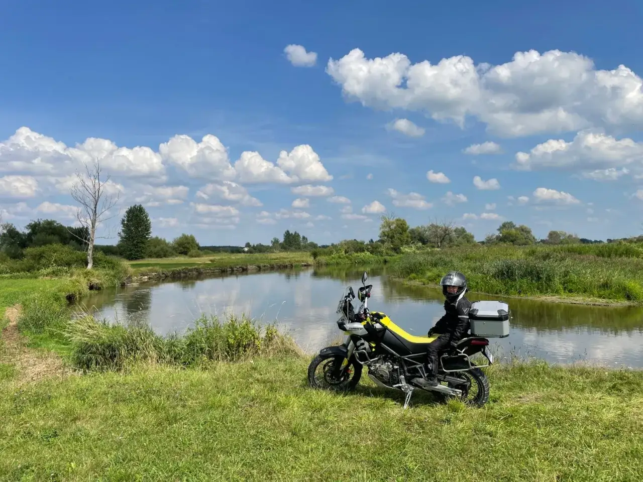 Motocyklista w kasku siedzi na motocyklu obok rzeki, na tle zielonej łąki i błękitnego nieba z chmurami.