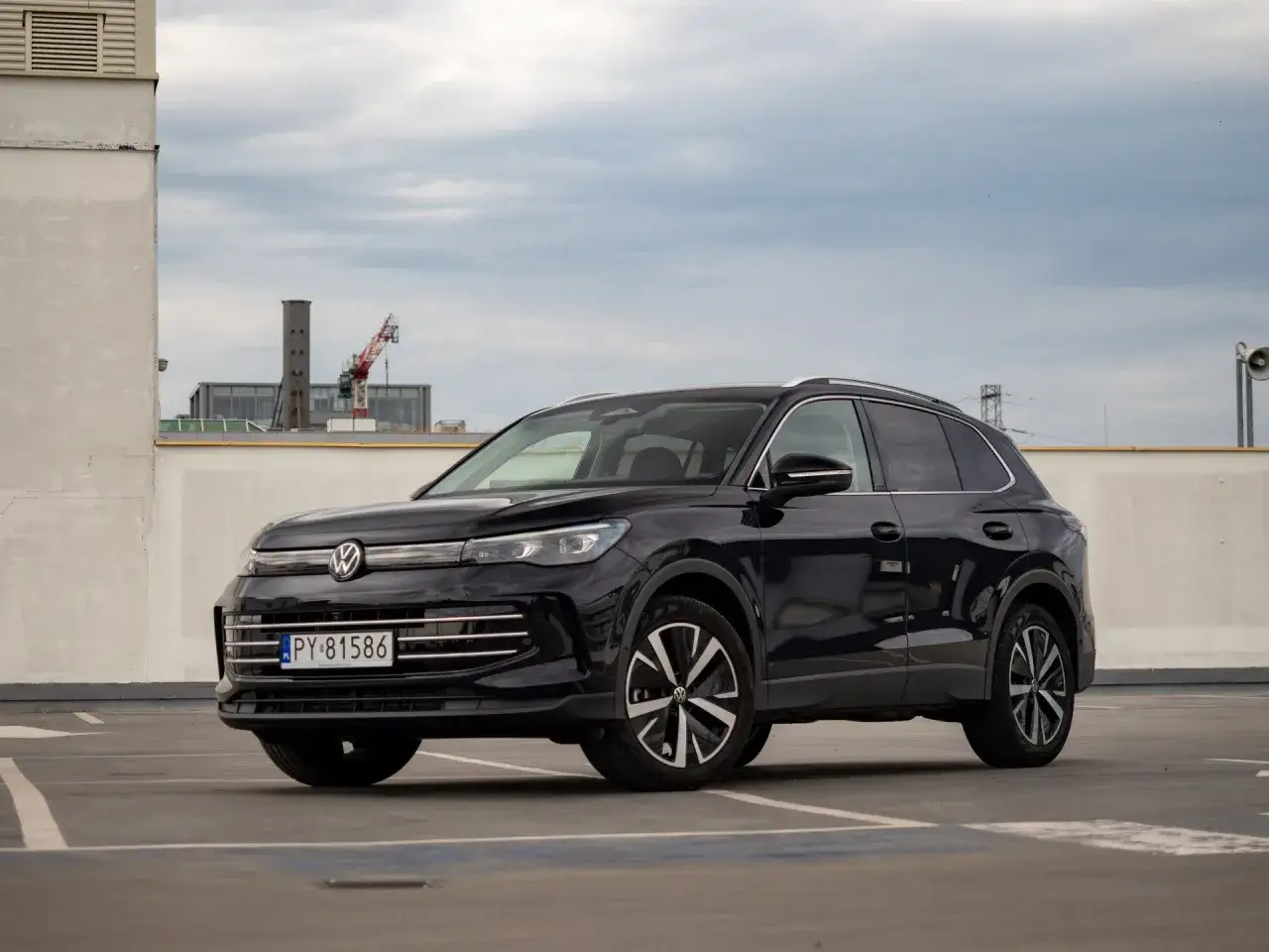 Czarny Volkswagen Tiguan na parkingu. W tle budynki i dźwig. Wiele osób szuka volkswagen tiguan opinie przed zakupem.