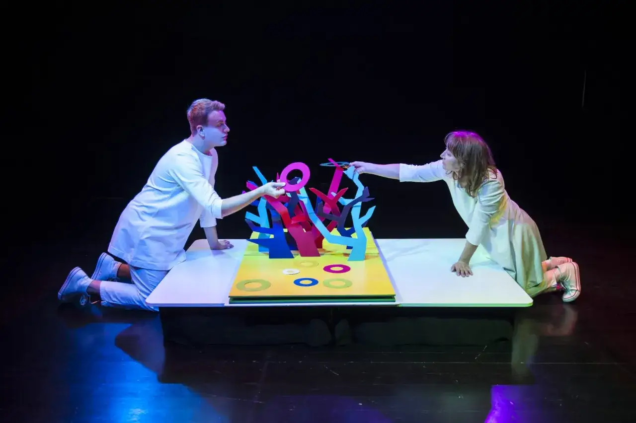 Dwoje aktorów w białych strojach tworzy magiczny świat na scenie. To teatr bajka Warszawa, gdzie kolorowe drzewka i pierścienie ożywają.