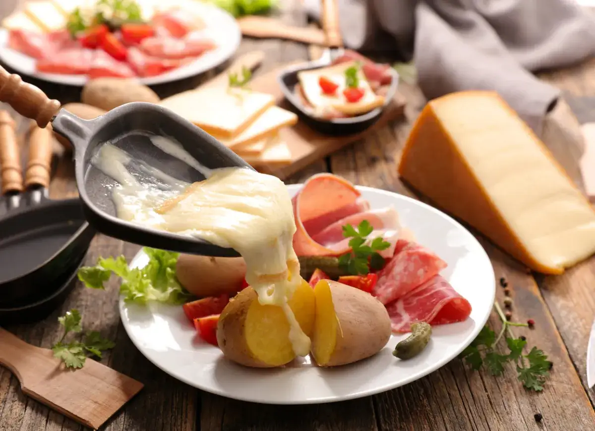 Roztopiony ser raclette spływa na ziemniaki i wędliny. Obok kawałek sera i talerz z dodatkami.