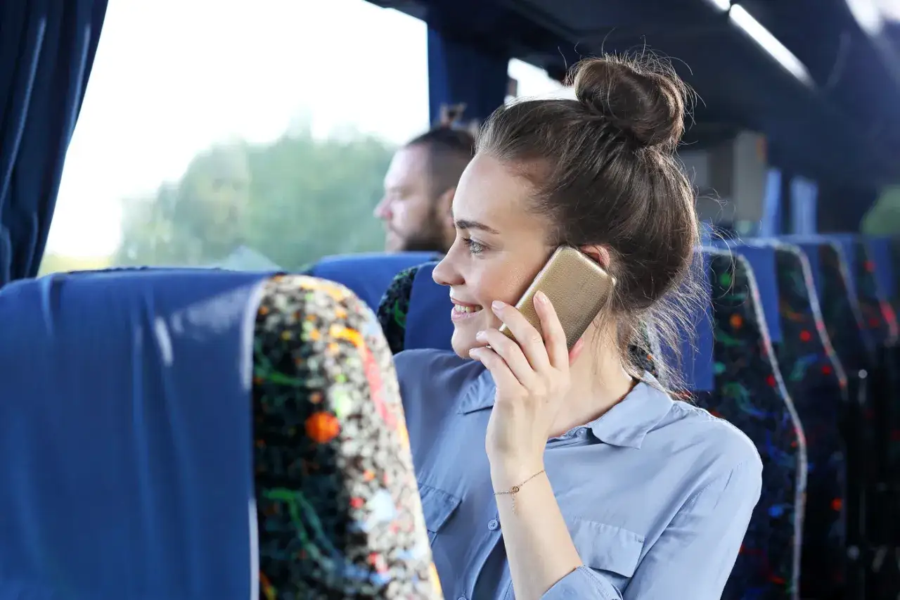 Uśmiechnięta kobieta rozmawia przez telefon w autobusie, szukając informacji, jak włączyć roaming Plus, by być w kontakcie podczas podróży.