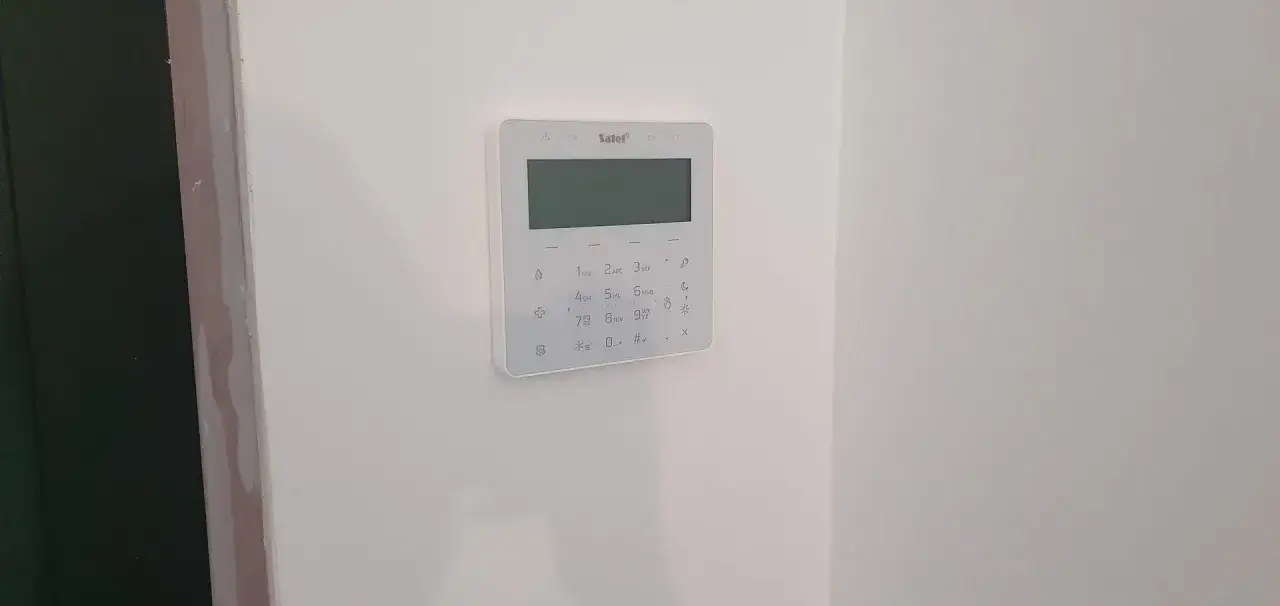 Biały panel alarmowy z klawiaturą numeryczną i ekranem na białej ścianie.
