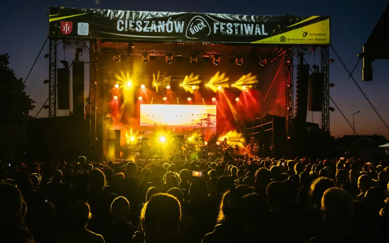 Tłumy bawią się na Cieszanów Rock Festiwal. Jasne światła sceny oświetlają zespół grający na tle zachodzącego słońca.