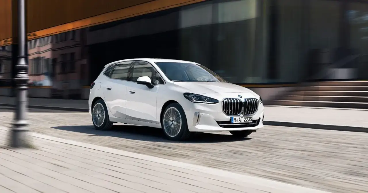 Biały BMW 2 Active Tourer w ruchu miejskim, z rozmytym tłem budynków i ulicy.