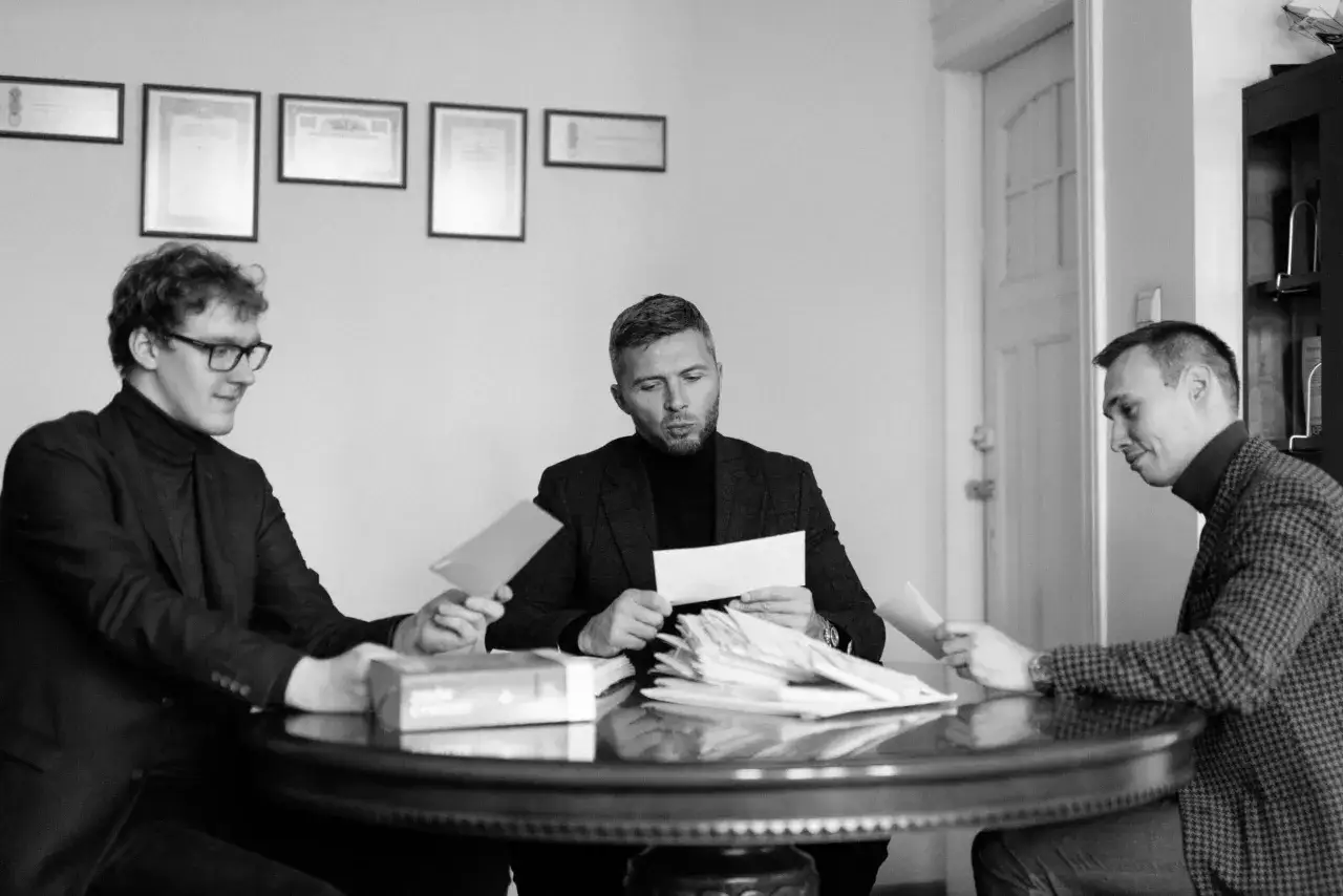 Trzech mężczyzn przy okrągłym stole analizuje dokumenty. Jeden z nich trzyma kopertę, być może dotyczącą alimentów.
