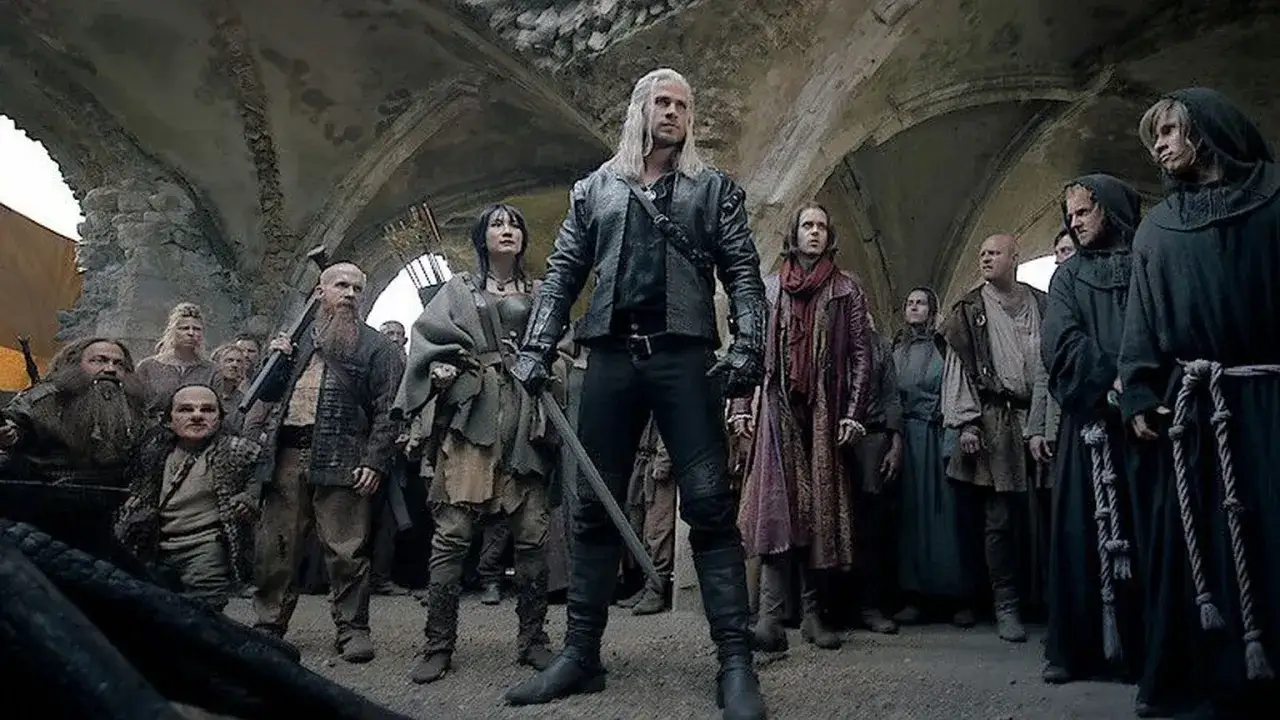 Geralt z Rivii stoi pośród tłumu, gdy ścieżki przeznaczenia w Wiedźminie splatają się. Widać krasnoludy, ludzi i zakapturzonych mnichów.