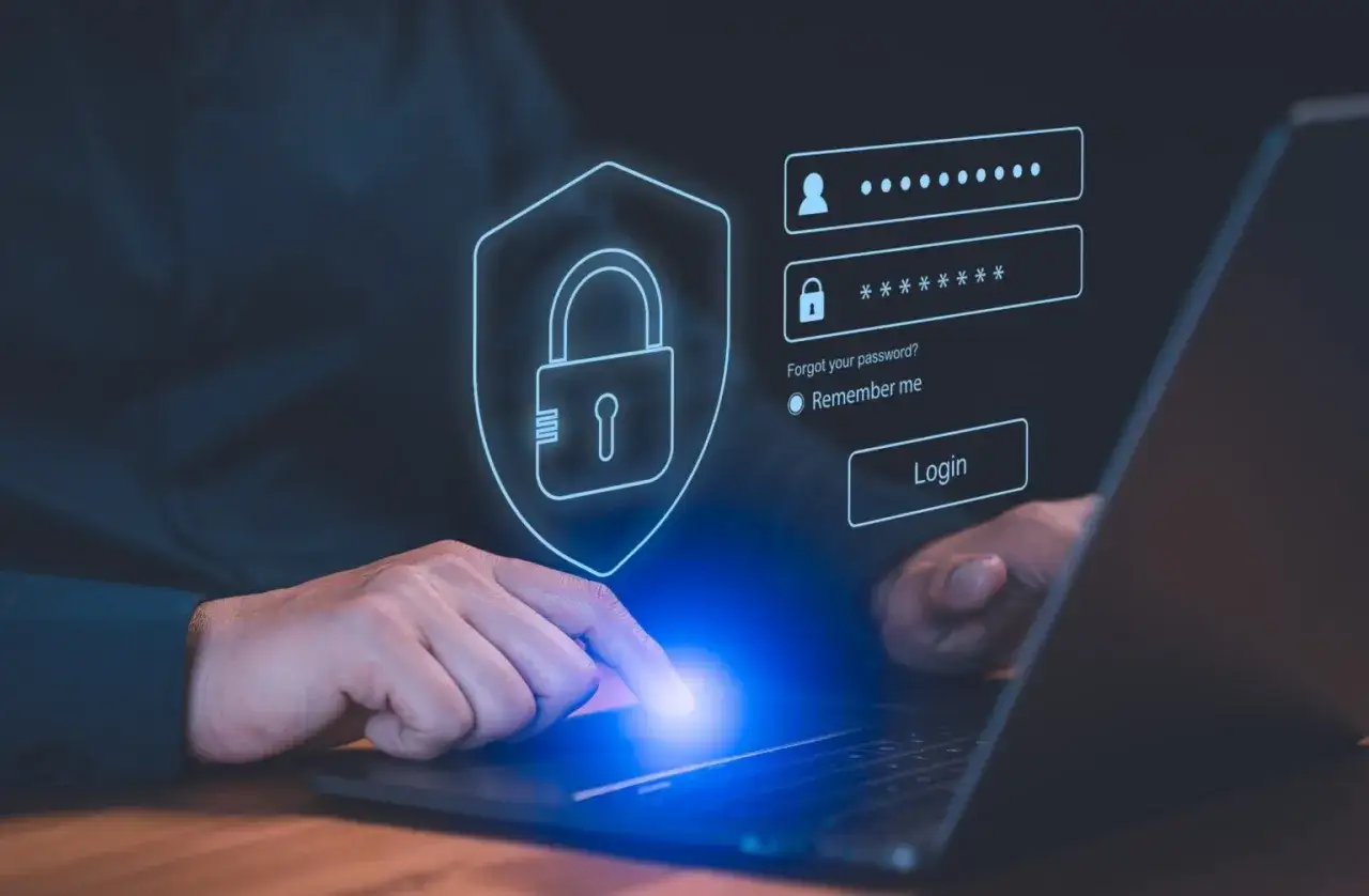 Dłoń dotyka ekranu laptopa z interfejsem logowania. Ikona tarczy z kłódką symbolizuje bezpieczeństwo, a obok pola na login i hasło znajduje się przycisk "Login". To nowoczesny menedżer haseł.