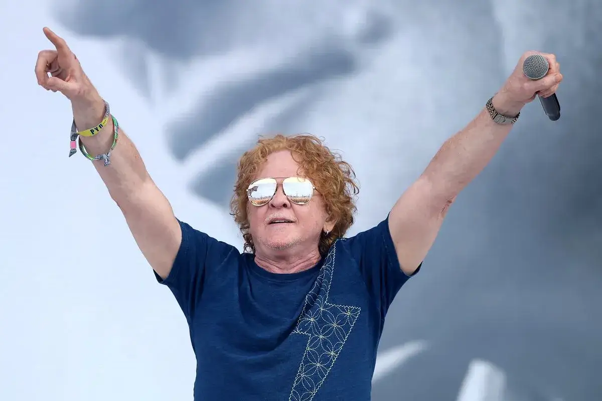 Wokalista Simply Red z uniesionymi rękami, trzymający mikrofon. Ma charakterystyczne rude włosy i okulary przeciwsłoneczne.