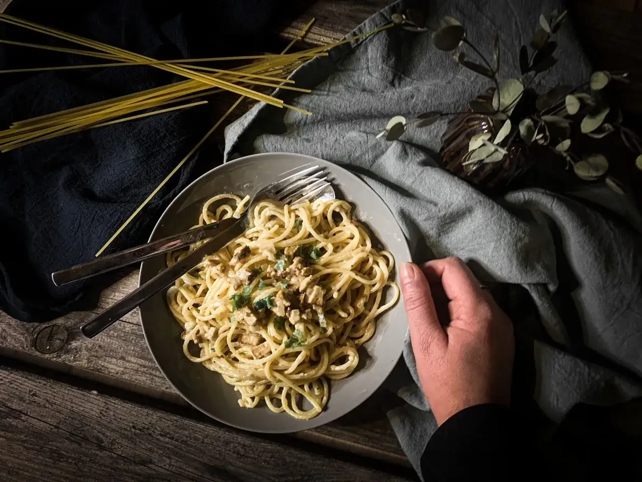 Teller mit cremiger, veganer Spaghetti Carbonara, garniert mit Kräutern. Daneben liegen ungekochte Spaghetti.
