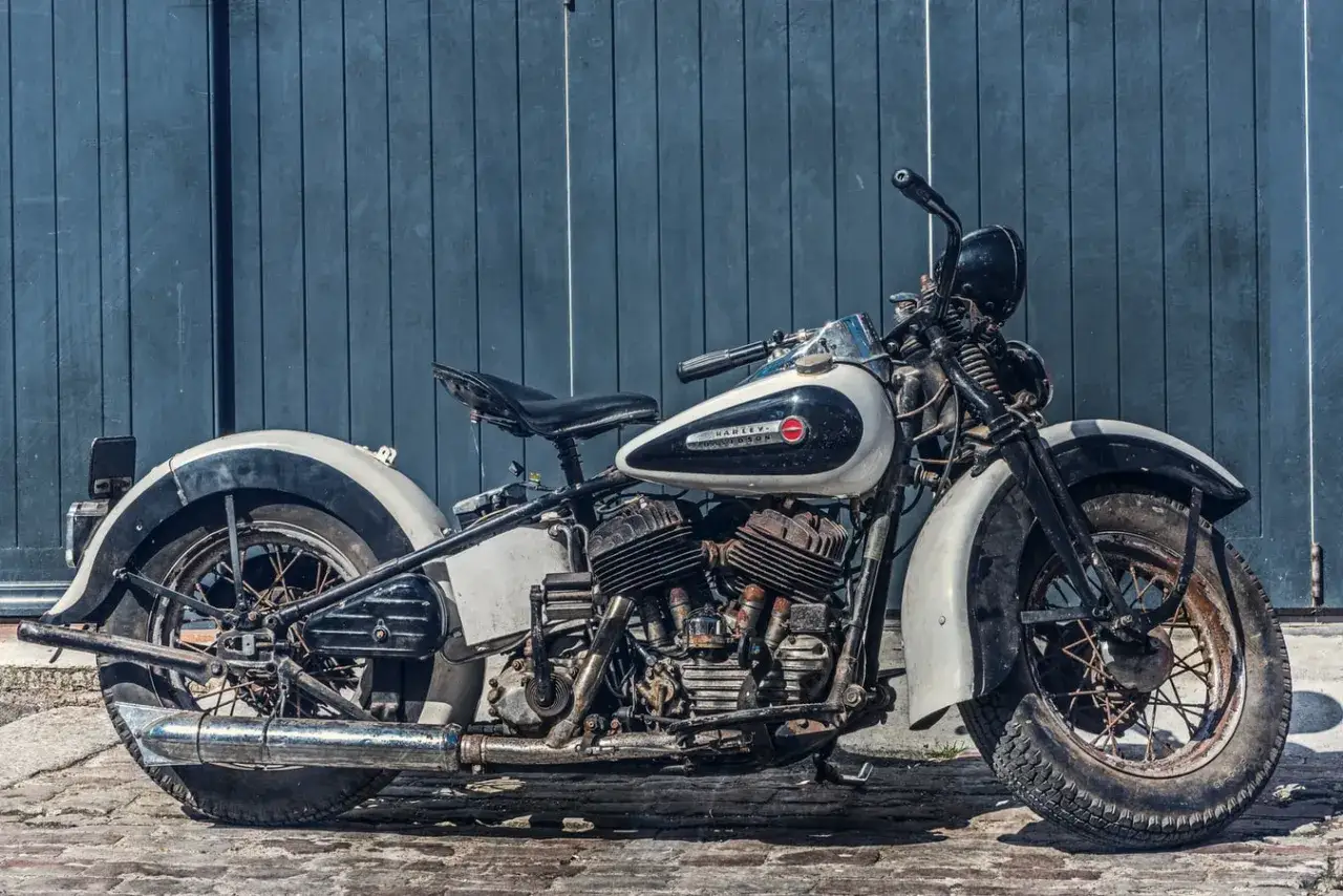 Stary motocykl Harley-Davidson w kolorze białym i czarnym, stojący na bruku przed niebieską, drewnianą ścianą.