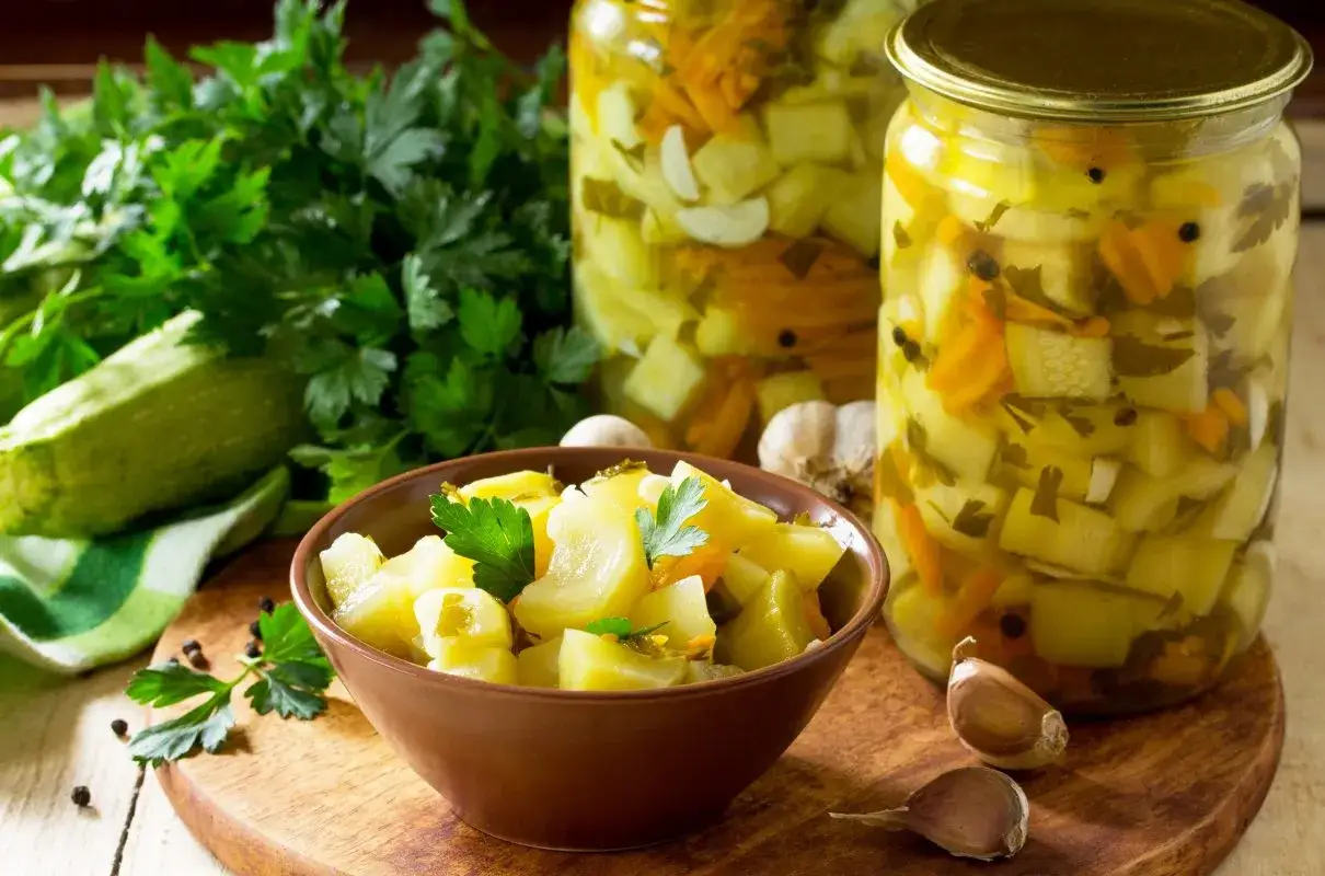 Słoiki z cukinią w zalewie słodko-kwaśnej z curry, miseczka z marynowaną cukinią, świeża cukinia, czosnek i natka pietruszki.