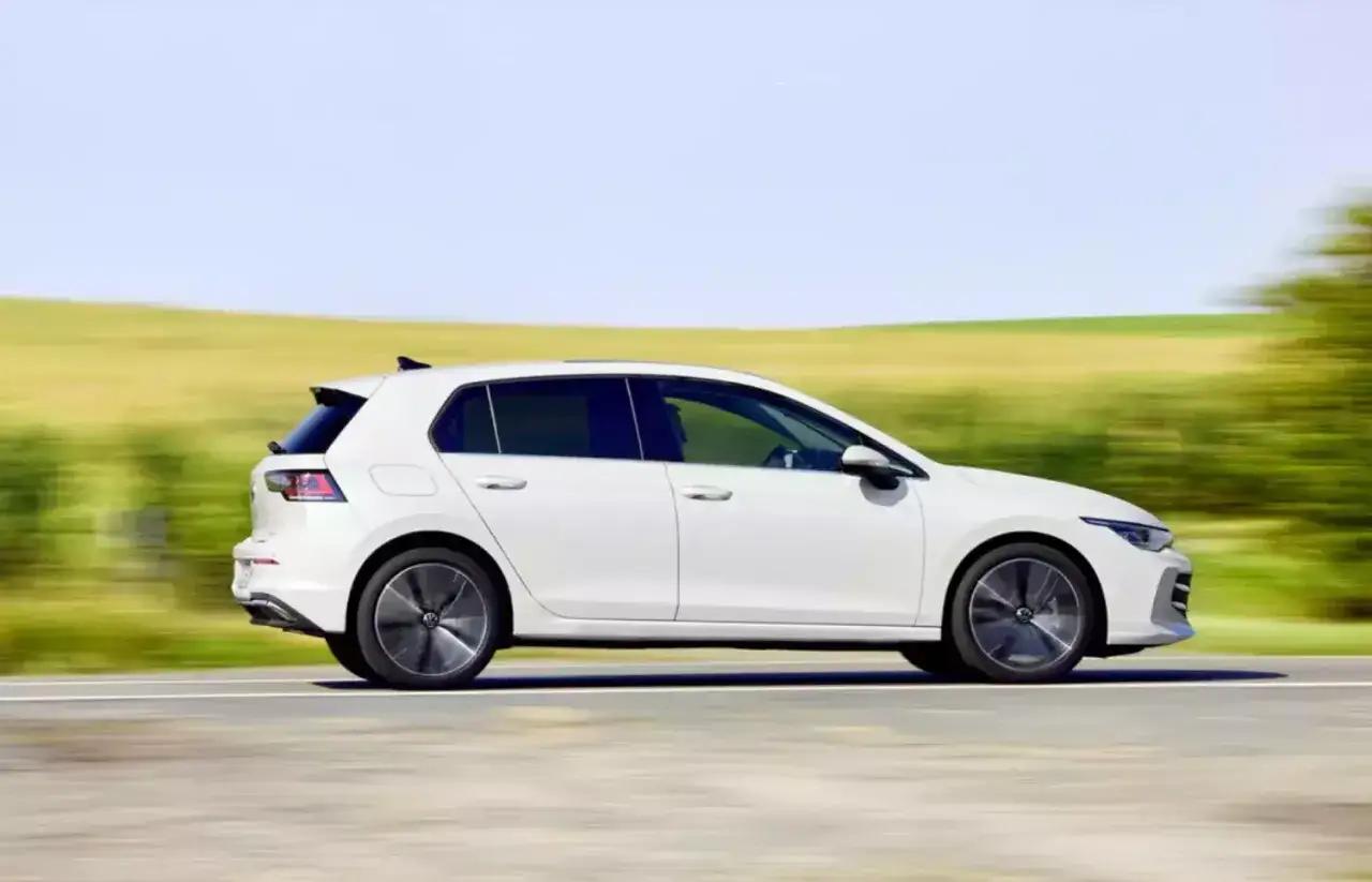 Biały hatchback pędzi po drodze, otoczony rozmytymi zielonymi polami i błękitnym niebem.
